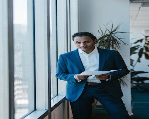 A focused professional man standing by a large window inside an office environment during a break